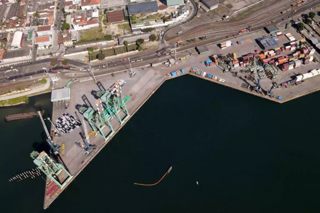 Vista aérea do porto com docas, contêineres de carga, áreas de transporte e infraestrutura portuária em um ambiente marítimo.