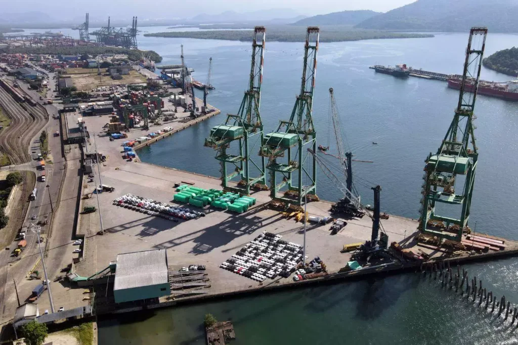 Imagem da área portuária industrial com grandes guindastes, embarcações no rio e infraestrutura para transporte de cargas, destacando o setor de logística e comércio marítimo.