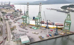 Vista panorâmica do porto com navio cargueiro e guindastes de movimentação de carga, destacando a atividade de transporte marítimo e logística no Brasil.