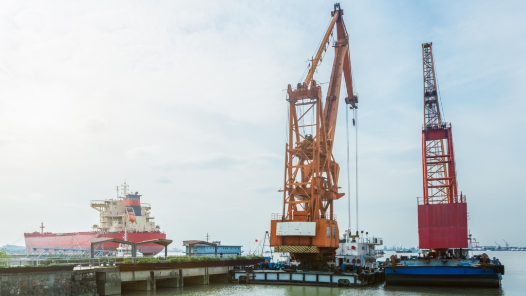 Imagem de um porto com guindastes carregando navios, destacando a operação de dragagem e movimentação de cargas marítimas, essencial para infraestrutura portuária.