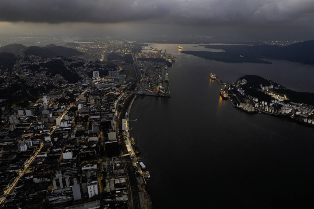 Vista aérea de Santos à noite com prédios iluminados, rios e montanhas ao fundo, sob céu nublado, destacando a beleza urbana e natural.