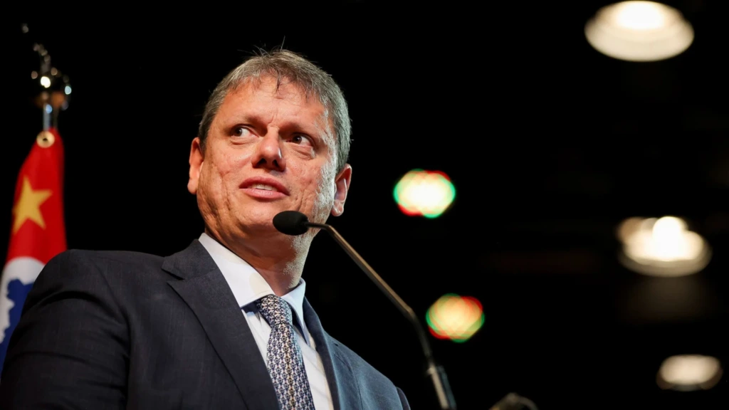 Imagem de Tarcísio de Freitas falando em um microfone em um evento, com fundo escuro e luzes ao fundo. Ele usa terno e gravata.