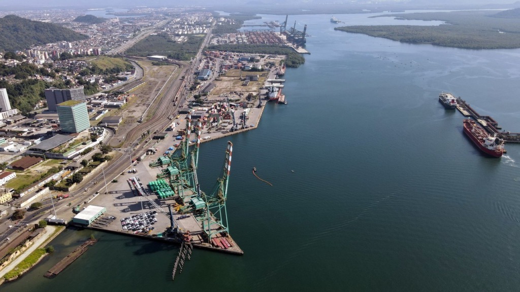 Imagem aérea de um porto no Brasil, com várias balsas, guindastes e estruturas de carga, ao lado de uma cidade costeira com edifícios e áreas industriais, vista de drone.
