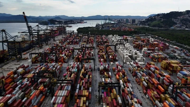 Imagem aérea do porto de Santos; pode-se ver múltiplos contêineres coloridos e guindastes de carga em atividade, sob um céu nublado.