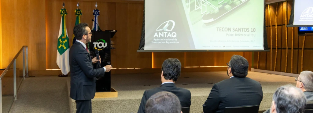 Representante do ANTAC apresentando o projeto TECON SANTOS 10 em reunião oficial, com detalhes técnicos exibidos na tela de projeção e bandeiras ao fundo.