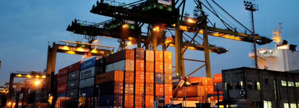 Imagem de um porto com contêineres de carga coloridos, guindastes e navios de transporte em operação, ilustrando logística portuária eficiente na exportação e importação.