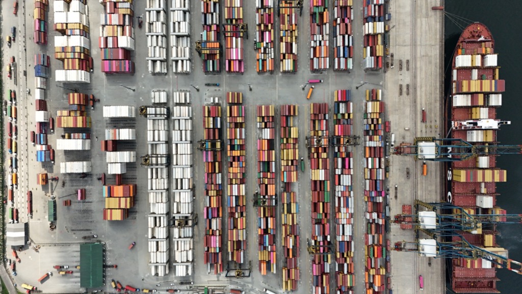 Imagem aérea de um porto de containers com várias pilhas coloridas de cargas e guindastes movimentando os containers, destacando a logística e o transporte marítimo.