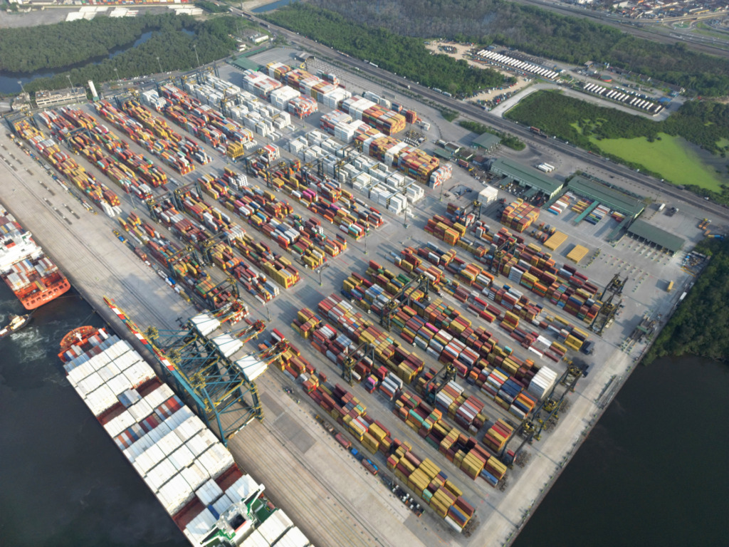 Imagem aérea de um porto de carga com diversos contêineres coloridos e navios atracados ao lado, situado às margens de um rio ou mar.