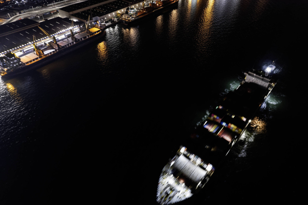 Imagem aérea noturna de um porto com navios atracados e iluminação refletindo na água, capturada com drone.