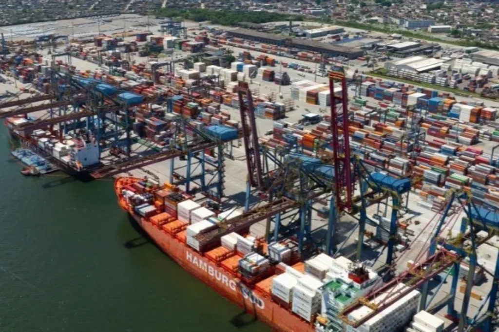 Imagem de um porto de contêineres com guindastes trabalhando na carga e descarga de barcos, ambiente de comércio internacional e logística.